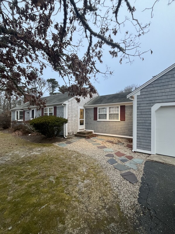 A residential property with a gravel driveway, a front yard with a small bush, and a house with a grey siding and red shutters.