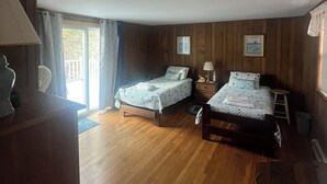 A room with two beds, wooden flooring, a desk with a lamp, and a window with curtains.