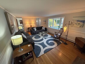 A spacious living room with a large sofa, a coffee table, and a patterned rug. There is a wooden floor, a rocking chair, and a lamp.