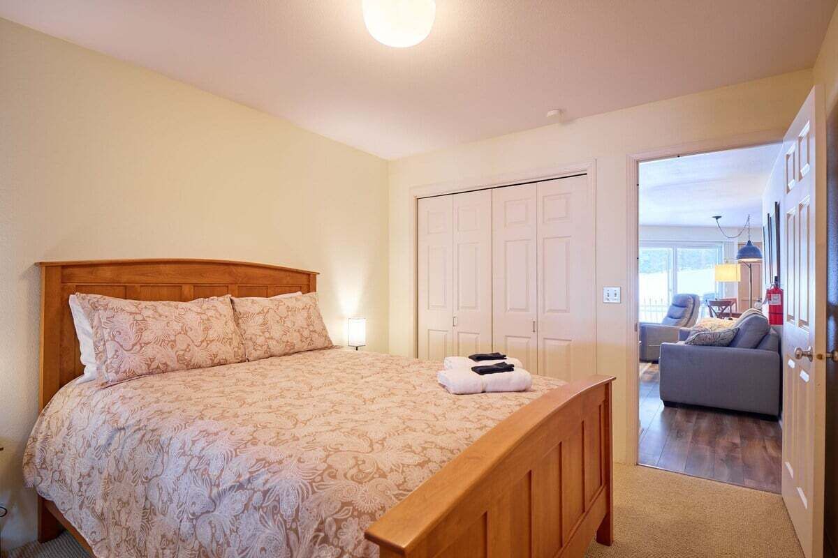 A bedroom with a wooden bed, a closet, and a view into a living area with a sofa and a lamp.