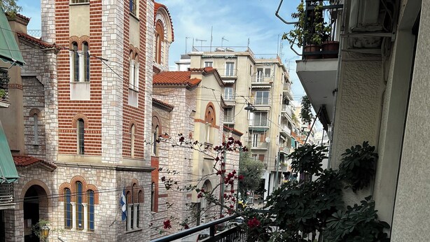 A balcony with a wooden table and chairs, potted plants, and a view of a street with cars and buildings. - Pnyka Apartment 2 (Athens)