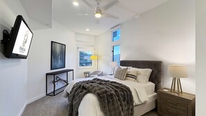 Un dormitorio con cama, mesita de noche, televisor, ventana y ventilador de techo.