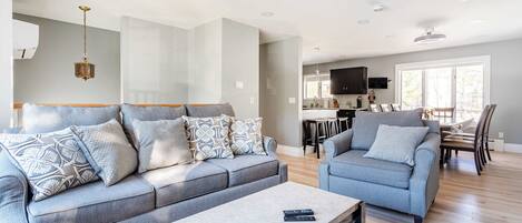 A modern living room with a grey sofa set, a coffee table, and a dining area with a TV and kitchen in the background.