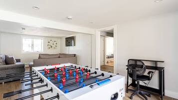 Een moderne woonkamer met een tafelvoetbalspel, een bank, een aan de muur bevestigde televisie en een bureau met stoel.