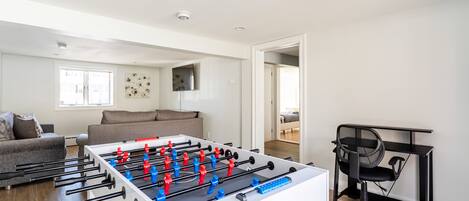 Una sala moderna con una mesa de futbolín, un sofá, un televisor montado en la pared y un escritorio con una silla.