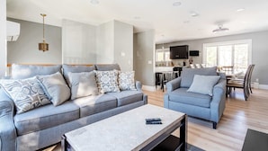 A modern living room with a grey sofa set, a coffee table, and a dining area with a TV and kitchen in the background. - The Woodland Escape | Pool Table | Fire Pit | 4 BD (Tobyhanna)