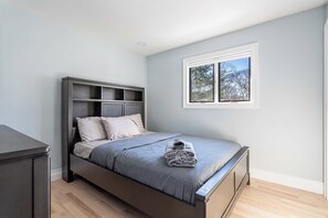 A bedroom with a bed, a nightstand, a window with a view of trees, and a wooden floor.