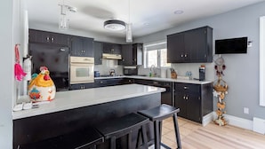A modern kitchen with dark cabinets, a large island with stools, a flat-screen TV, and a decorative plant. - The Woodland Escape | Pool Table | Fire Pit | 4 BD (Tobyhanna)