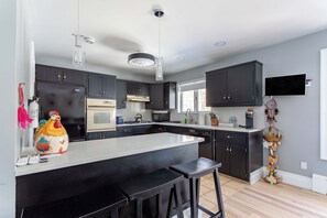 A modern kitchen with dark cabinets, a large island with stools, a flat-screen TV, and a decorative plant. - The Woodland Escape | Pool Table | Fire Pit | 4 BD (Tobyhanna)