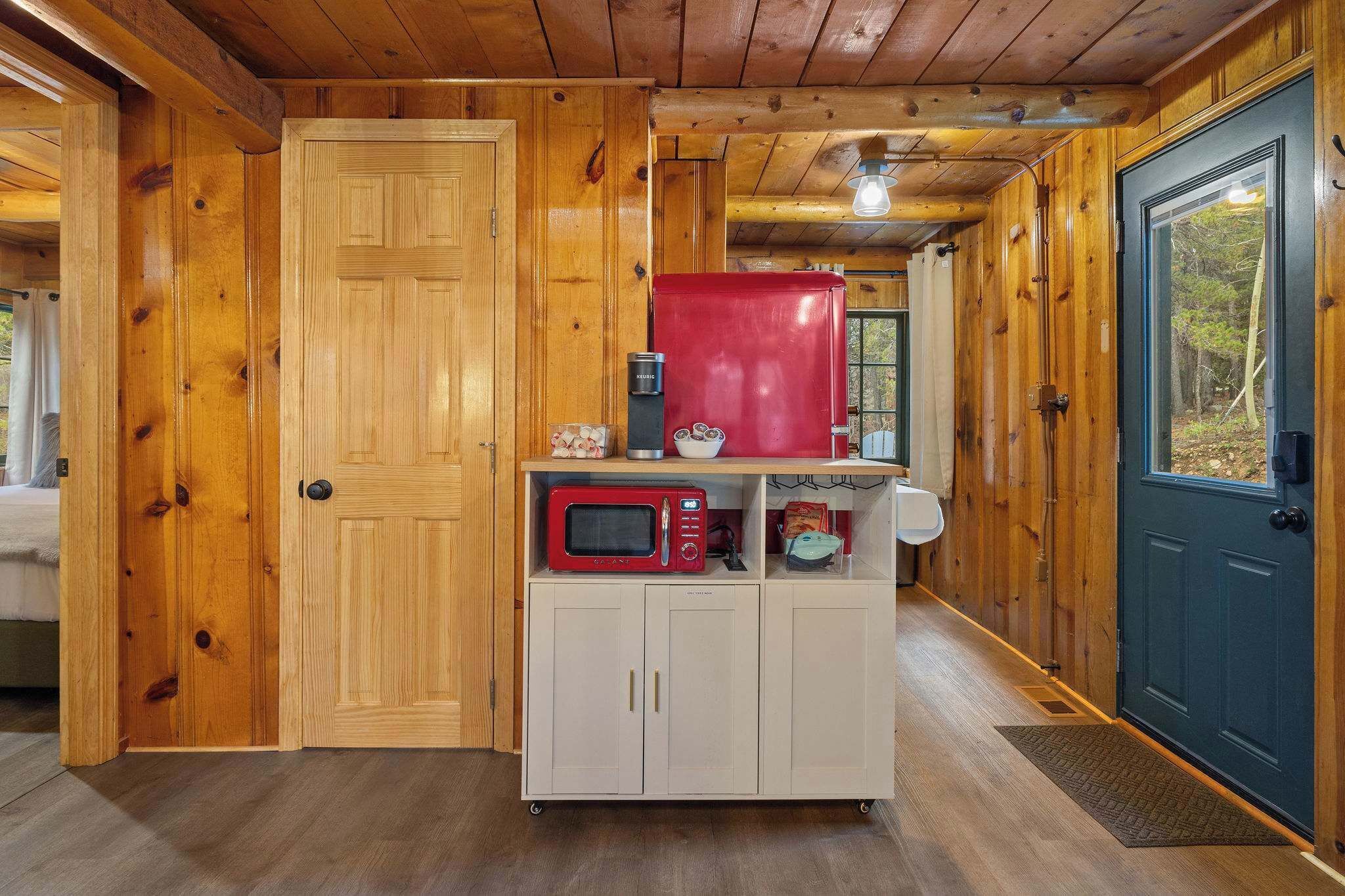 Cocina pequeña con paredes de madera y una puerta azul, que incluye un microondas y un refrigerador compacto.