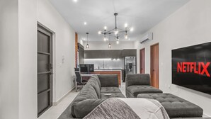 A modern living room with a large flat-screen TV, grey sofas, and a kitchen in the background.