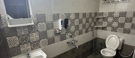 Une salle de bain avec un lavabo, des toilettes et une douche. On y trouve un miroir, un porte-serviettes et une poubelle bleue.