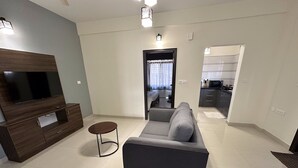 A modern living room with a flat-screen TV, dark wood entertainment unit, grey sofa, and a small round table. - Trinetra Homes 61 (Bengaluru)