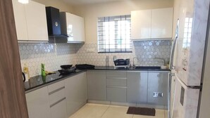 A modern kitchen with white cabinets, a black countertop, and a stainless steel refrigerator. - Trinetra Homes 61 (Bengaluru)
