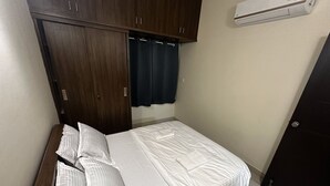 A single bed with white bedding, a dark wooden wardrobe, and a wall-mounted air conditioner. - Trinetra Homes 61 (Bengaluru)
