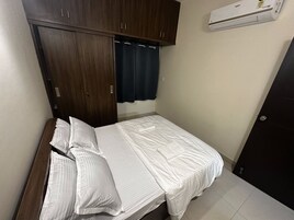 A single bed with white bedding, a dark wooden wardrobe, and a wall-mounted air conditioner.