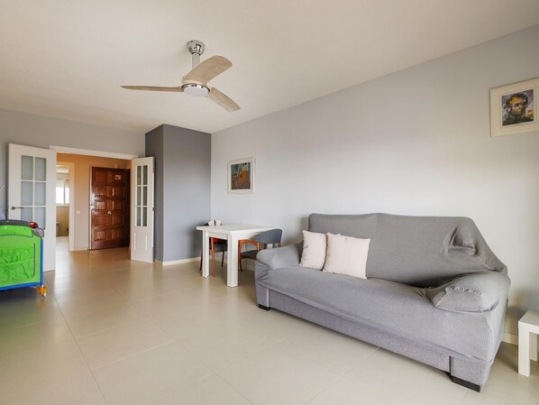 Una sala de estar con un sofá gris, una mesa blanca, un ventilador de techo y un corralito verde.
