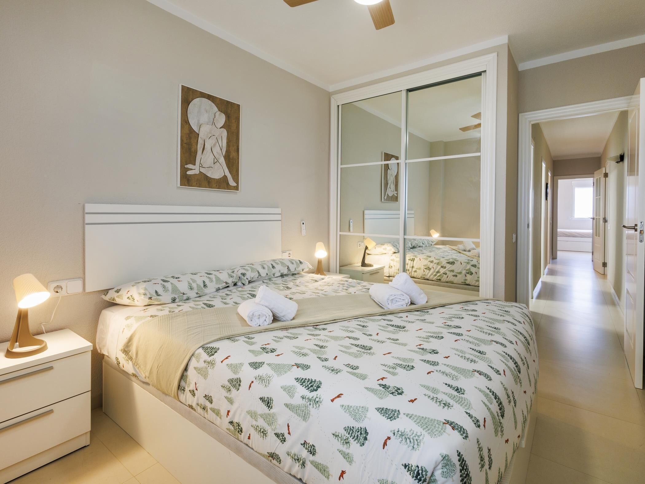 Een hotelkamer met een groot bed, een nachtkastje met een lamp, een spiegel en een schilderij aan de muur.