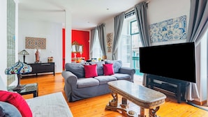 A living room with a grey sofa, red and pink cushions, a wooden coffee table, a TV, and a large window with curtains.