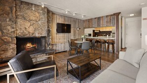 Un salon chaleureux avec une cheminée en pierre, un coin repas et une cuisine en arrière-plan.