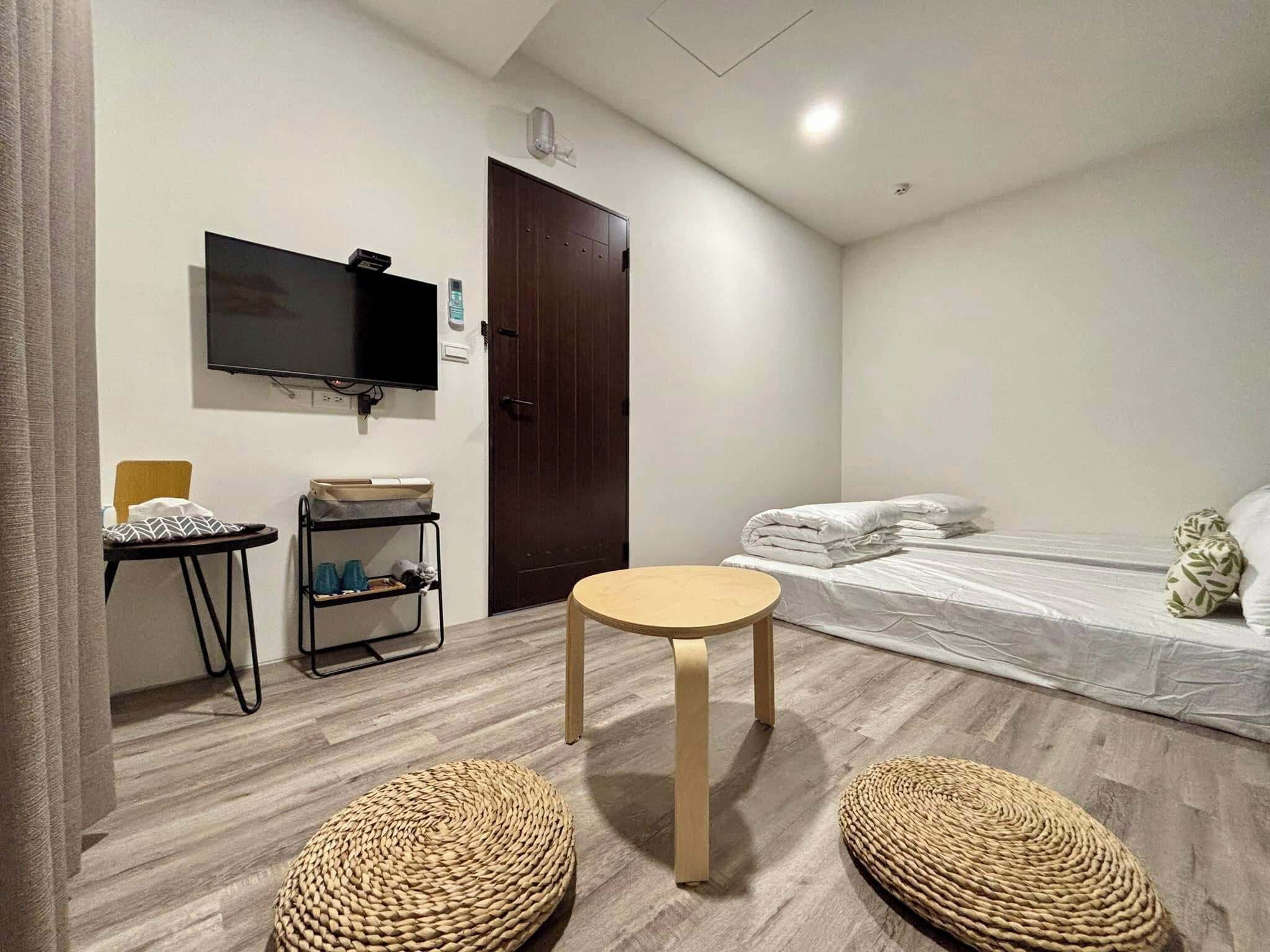 Habitación de hotel con una cama, una mesita redonda pequeña, un televisor montado en la pared, una puerta de madera y un estante con objetos.