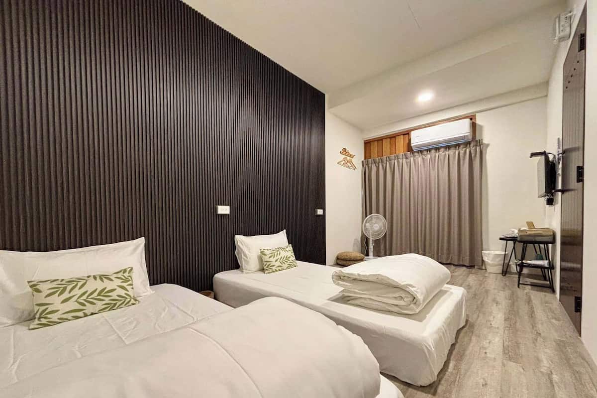 A hotel room with two beds, a small table, a fan, and a window with curtains.