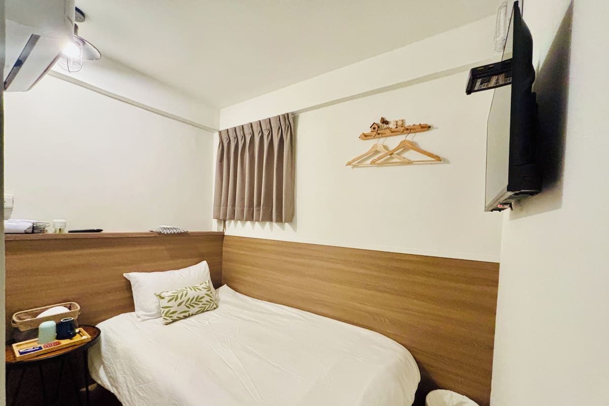 A hotel room with a wooden headboard, a bed with white bedding, a bedside table with a lamp, a small table with toiletries, and a TV mounted on the wall.