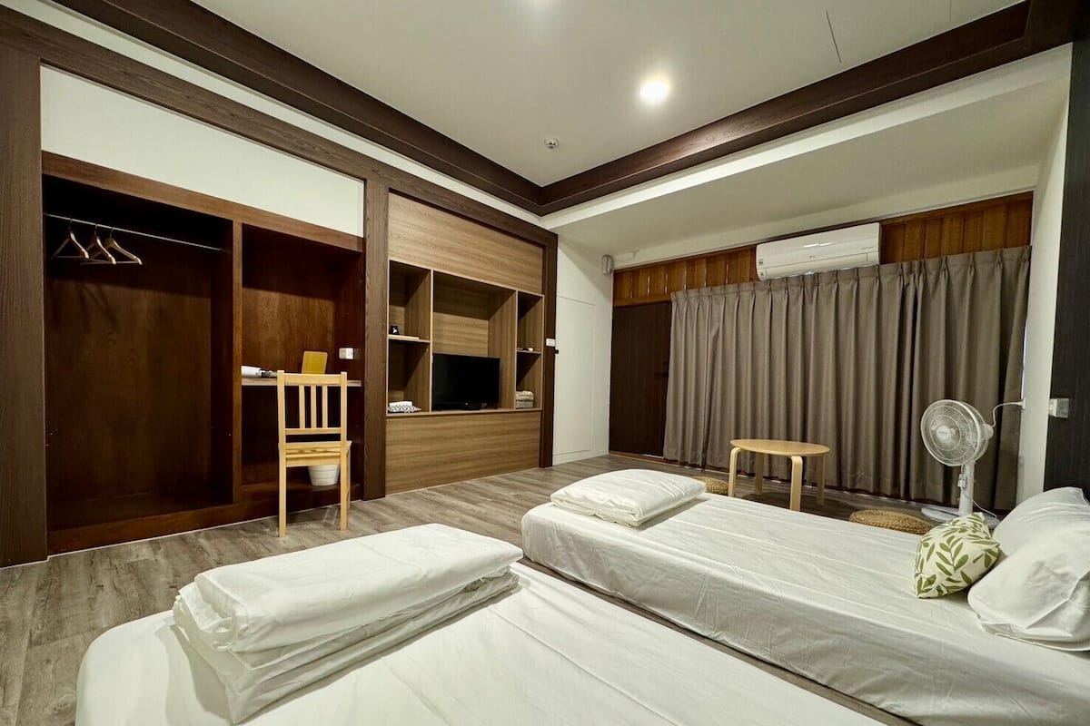 A hotel room with two single beds, a wooden wardrobe, a desk with a chair, a television, and a fan.
