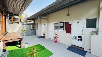 An outdoor area with a wooden table, a green artificial turf, a potted plant, a fire extinguisher, and a door with shoe storage.