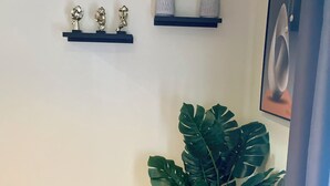 A corner of a room with a wooden floor, a large potted plant, two wall-mounted shelves with decorative items, and a framed picture on the wall.