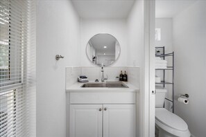 A bathroom with a white sink, a round mirror, a toilet, and a shelf with toiletries. - Cozy studio in Downtown Austin! (Austin)