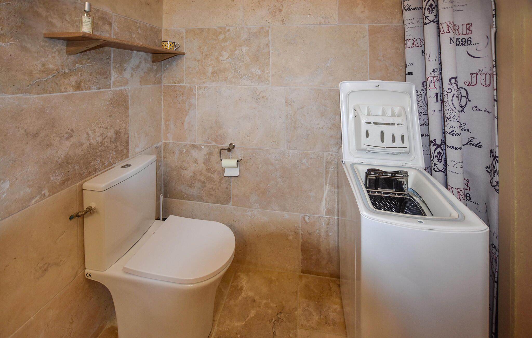 A compact bathroom with a toilet, a washing machine, and a shelf with a bottle and a mug.