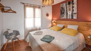 Ein Schlafzimmer mit Bett, Stuhl, einem Fenster mit Vorhängen, einem Wandbild und einem Holz-Kopfteil.