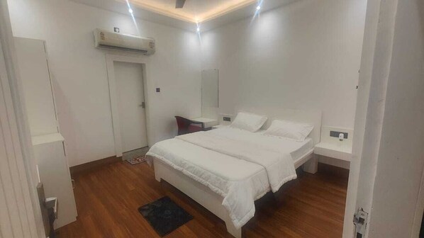 A bedroom with a bed, bedside tables, a wardrobe, an air conditioning unit, and a door. - Sunrise Meadows Hotel Praba's (Varanasi)