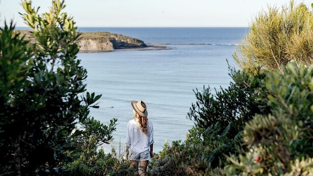 Una persona está de pie en un acantilado, contemplando un mar tranquilo con una isla rocosa al fondo.