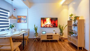 Een moderne woonkamer met een houten eettafel gedekt voor zes personen, een flatscreen televisie waarop een haard te zien is, en een houten boekenkast.