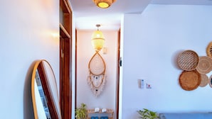 Een hal met een houten vloer, een hanglamp, een spiegel, een aan de muur bevestigd dromenvanger en een blauwe bank met een bloempot.