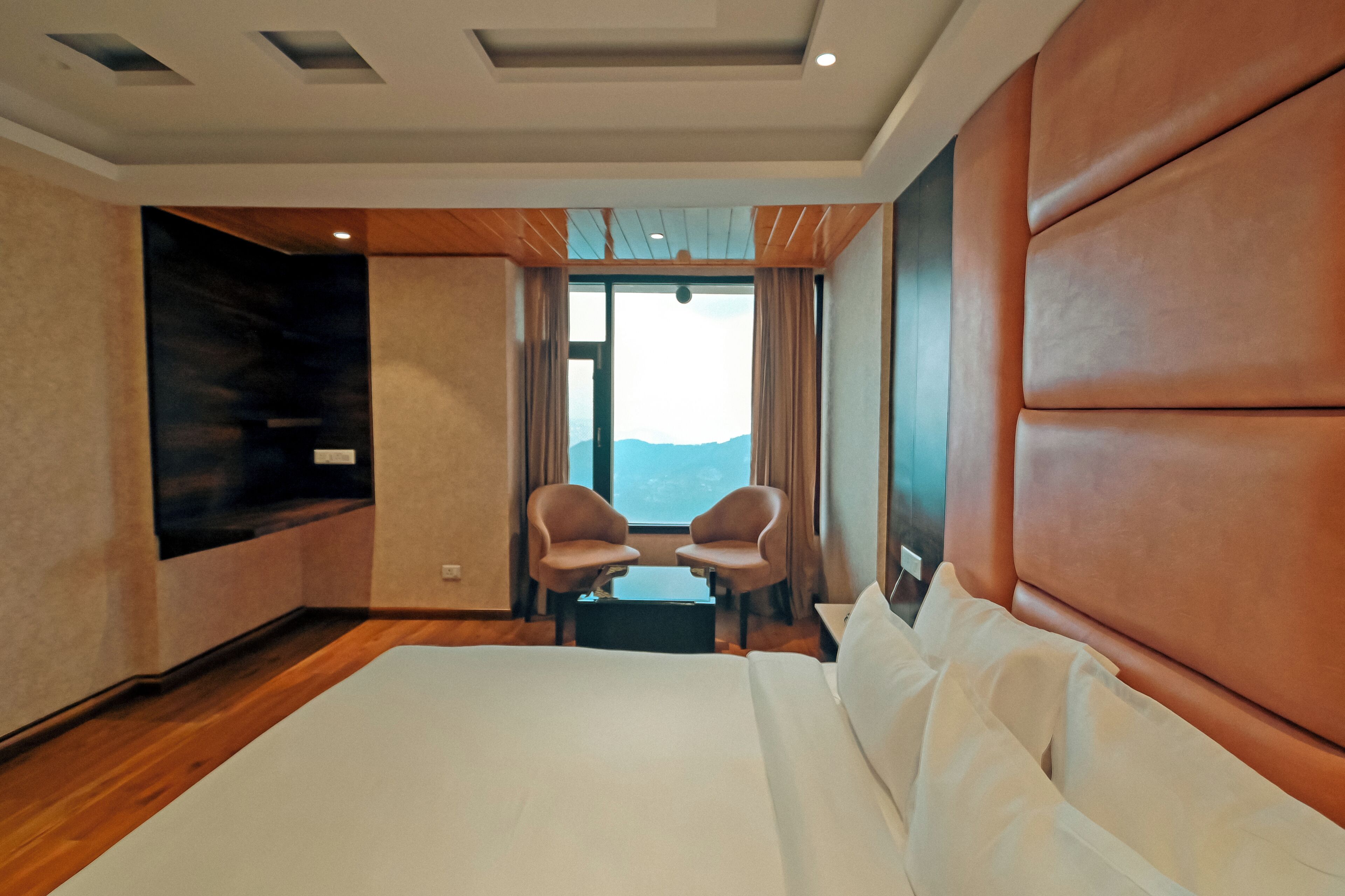 Quarto de hotel com uma cama grande, televisão, duas cadeiras e vista para as montanhas.
