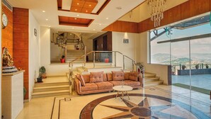 A spacious living area with a large sofa, a coffee table, and a decorative floor design. A staircase leads to an upper level with a view of the outdoors. - Alpine Holiday Inn, Kufri, Shimla (Theog)