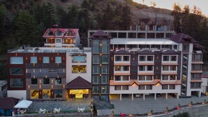 A multi-story hotel with a mix of modern and traditional architecture, featuring balconies, large windows, and a central courtyard with outdoor seating. - Alpine Holiday Inn, Kufri, Shimla (Theog)