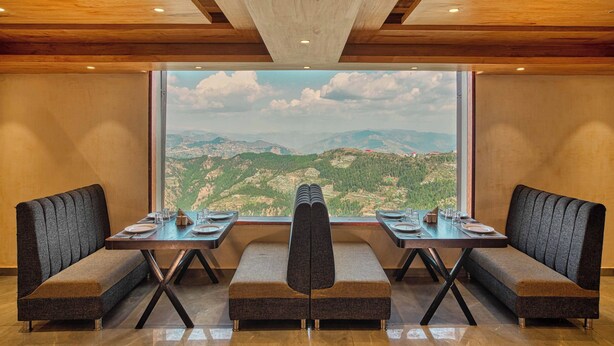 A dining area with a table set for four, a bench in the center, and a large window offering a mountain view. - Alpine Holiday Inn, Kufri, Shimla (Theog)