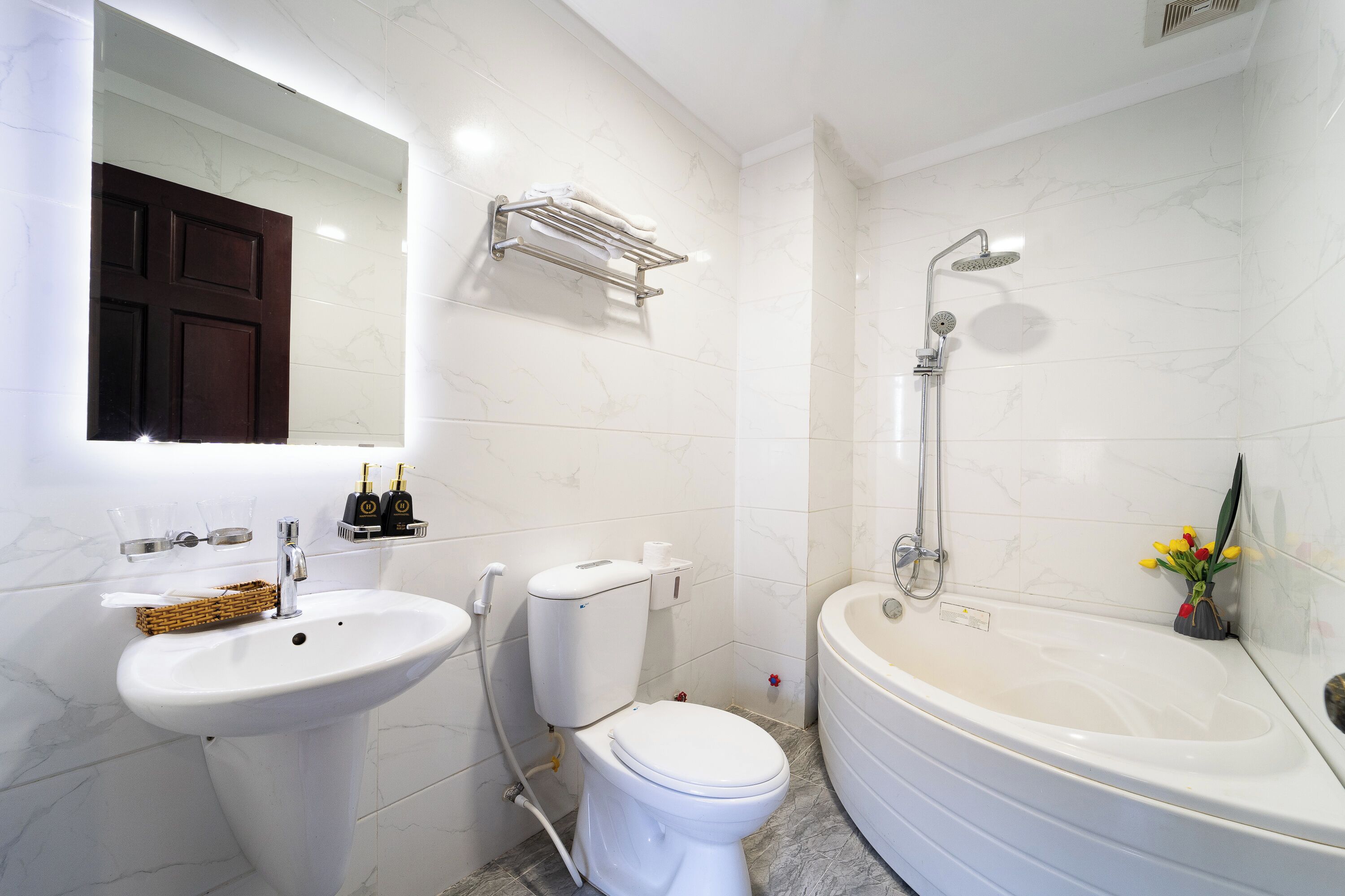 A bathroom with a sink, toilet, and bathtub. There is a mirror above the sink, a towel rack, and a small shelf with bottles.