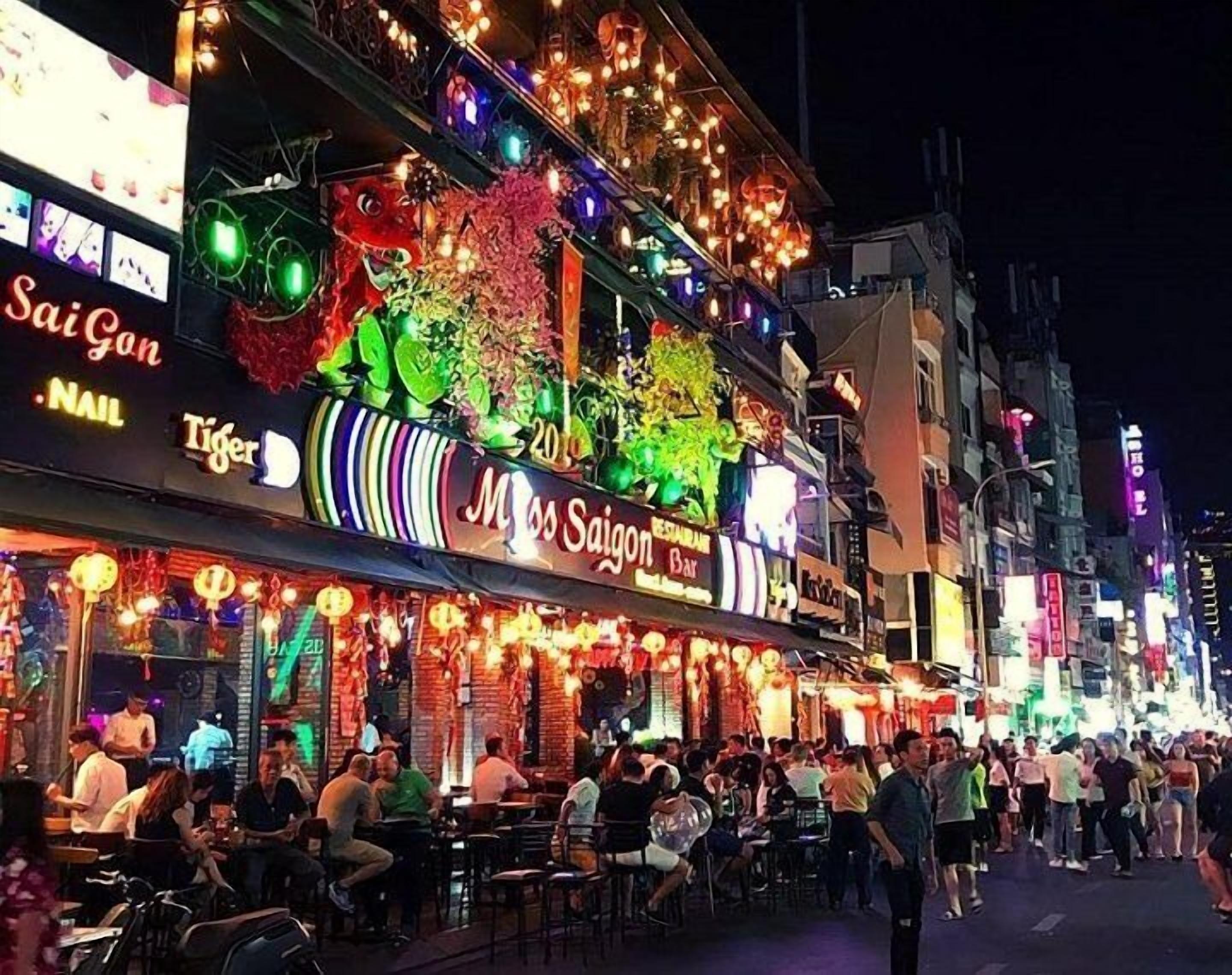 A bustling street scene with a brightly lit restaurant named "Miss Saigon Bar" featuring a colorful facade and outdoor seating.