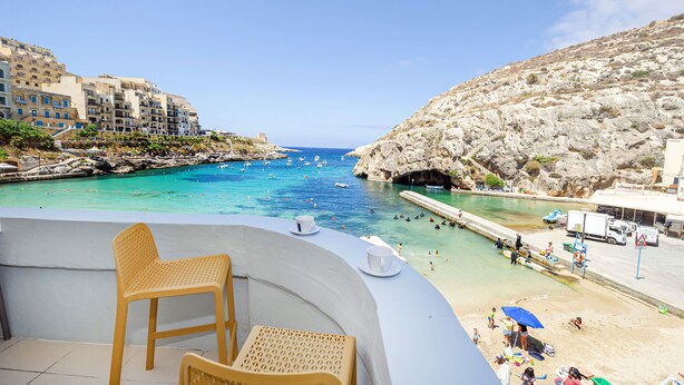 Un balcon agrémenté de chaises et d’une table en osier, offrant une vue sur une zone côtière avec des bâtiments, une colline rocheuse et une mer bleue et limpide où naviguent des bateaux.
