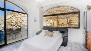 Une chambre dotée d’une grande fenêtre en arc de cercle, d’un lit, d’un bureau avec des chaises et offrant une vue sur un paysage rocheux et des bâtiments.
