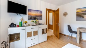 A modern living room with a flat-screen TV, a wooden cabinet with decorative items, a dining area with a table and chairs, and a door leading to another room. - Apartment 'Rumphorst' with Shared Terrace, Private Garden and Wi-Fi (Kirchhundem)