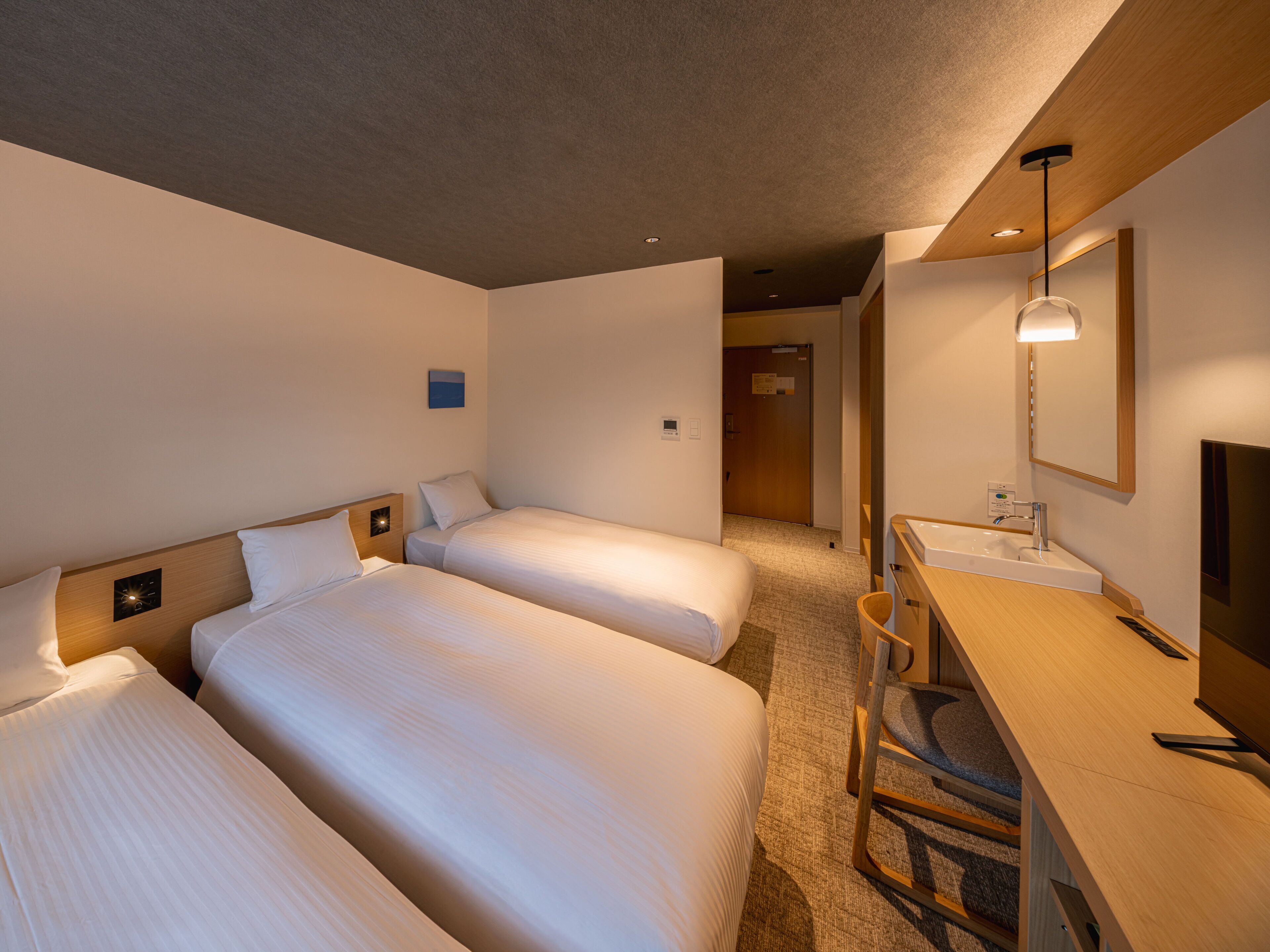 A hotel room with two beds, a desk with a chair, a mirror, and a small wall-mounted TV.