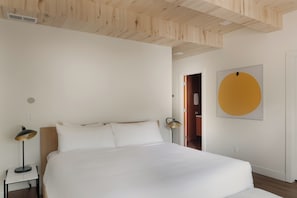 Une chambre avec un grand lit, une table de chevet avec une lampe, une porte donnant sur une autre pièce et un mur orné d’une œuvre d’art circulaire.