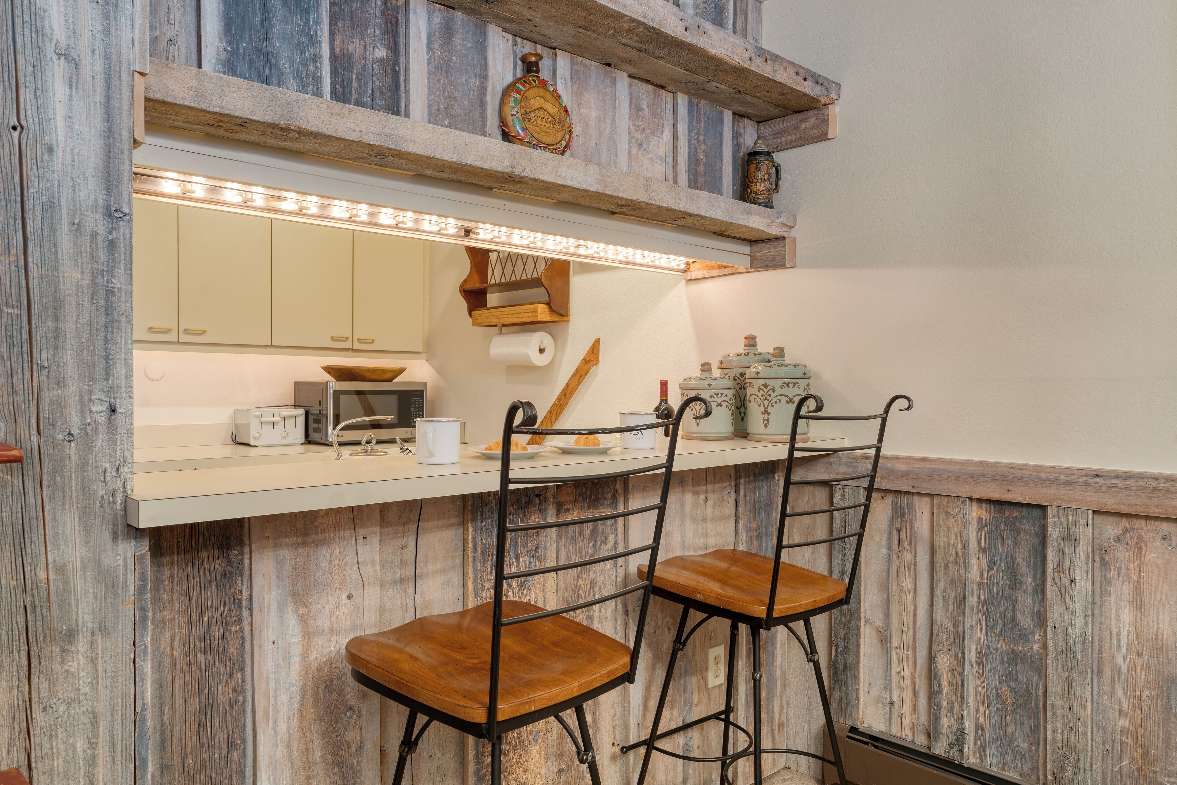 A kitchen with wooden paneling, a bar area with two stools, a countertop with a toaster, and a hanging shelf with decorative items.