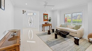 A spacious living room with a white sofa, a coffee table, and a large window with blinds. - Spacious 4BR Poolside Escape (Burleigh Waters)